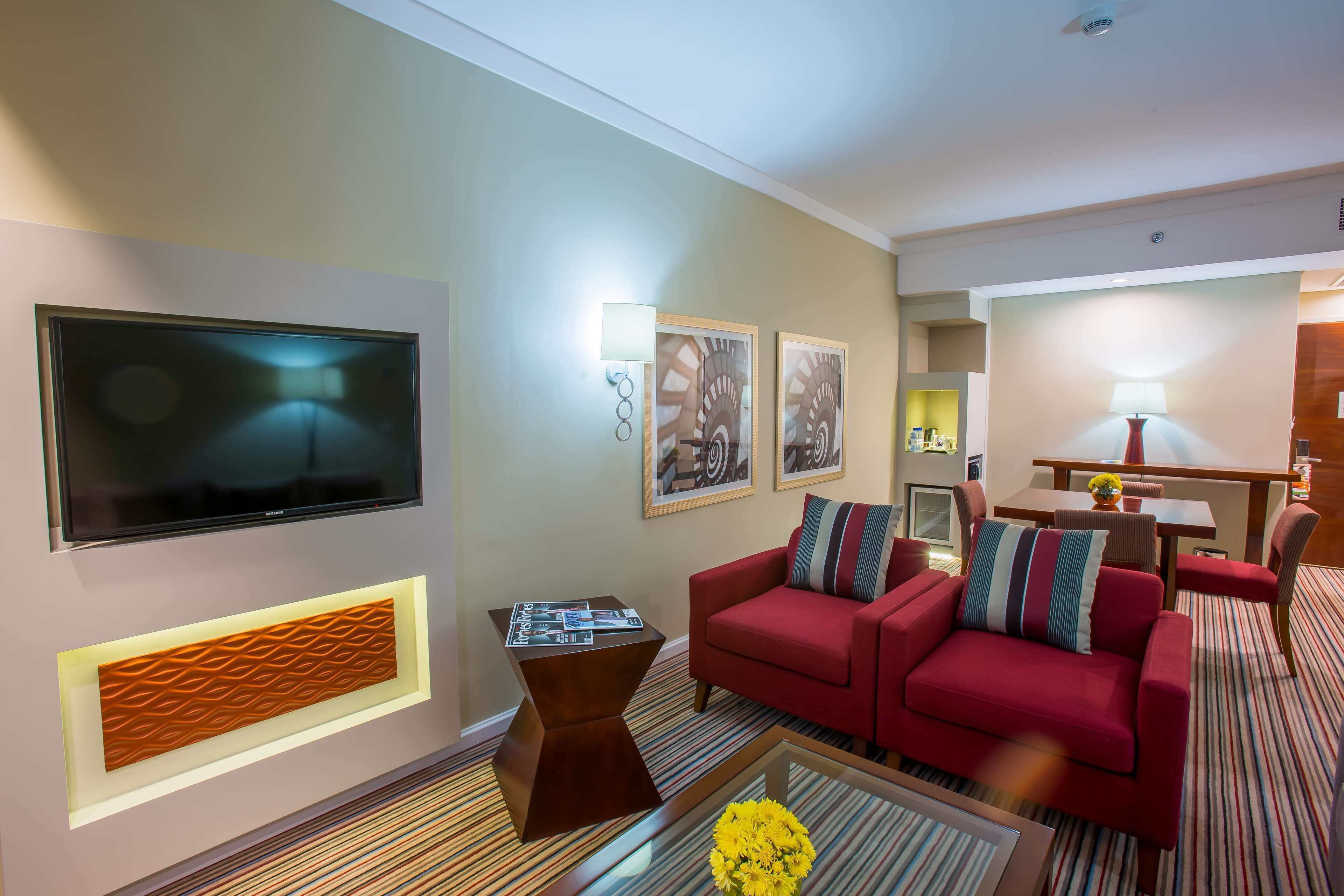 A modern hotel room with a red sofa, a flat-screen TV, framed artwork, and a patterned carpet.
