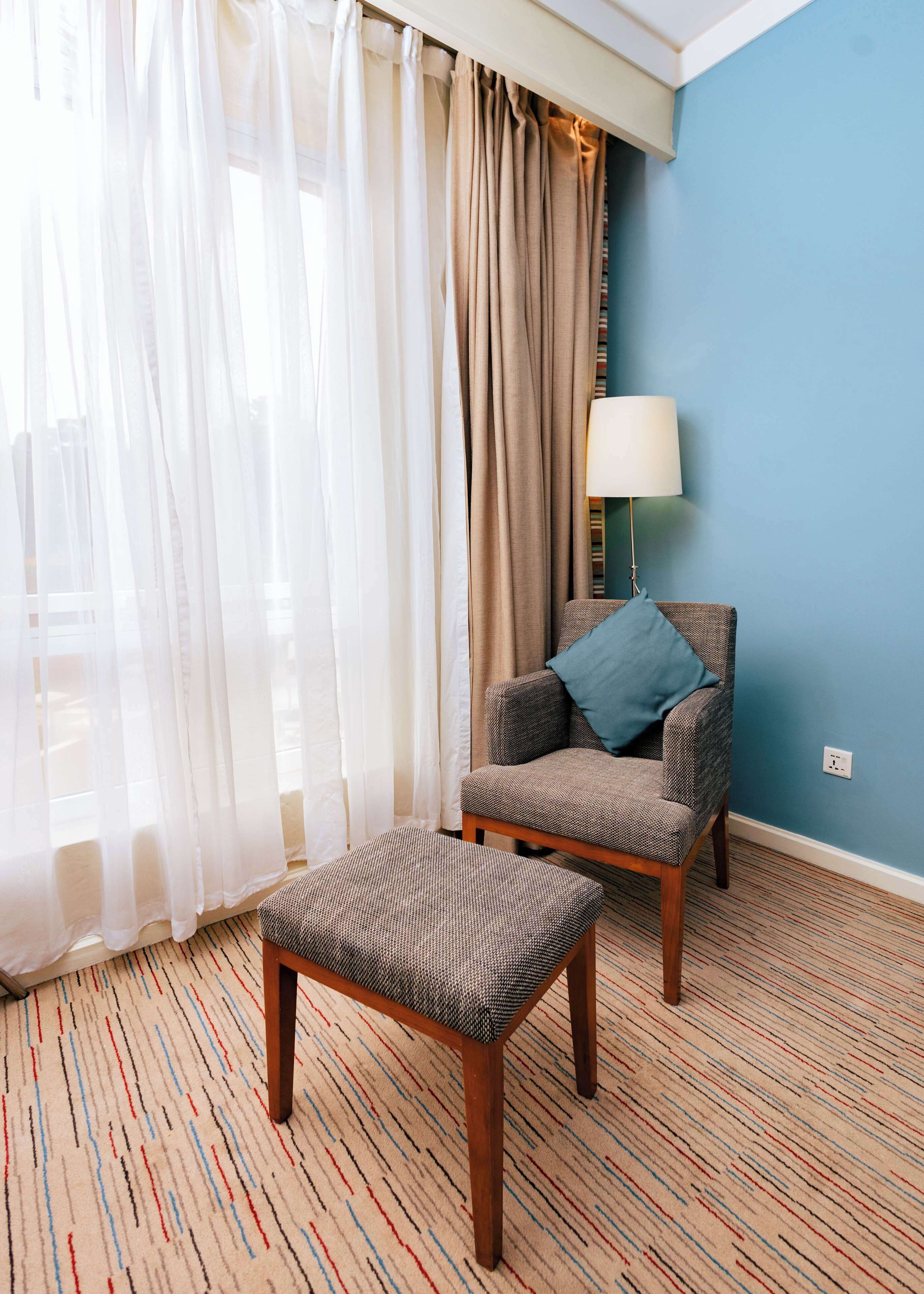 A room with a patterned carpet, a chair with a cushion, a small ottoman, and a lamp.