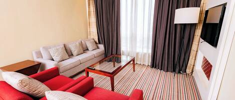 A hotel room with a red sofa, a beige sofa, a glass coffee table, a TV mounted on the wall, and a striped carpet.
