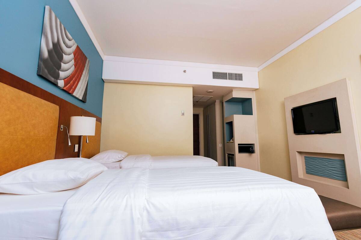A hotel room with a large bed, a flat-screen TV mounted on the wall, a ceiling vent, and a wall-mounted light fixture.