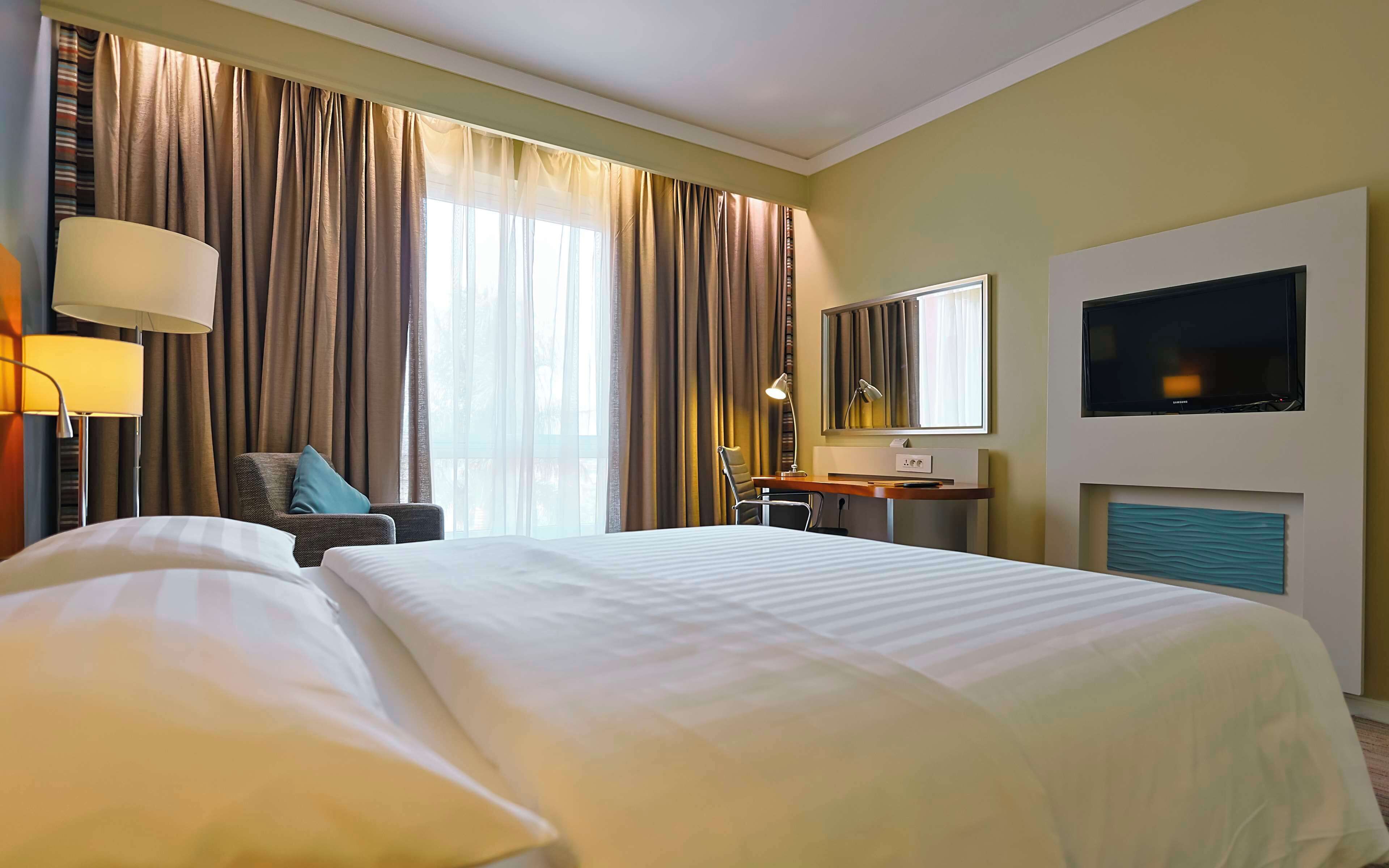 A hotel room with a large bed, a desk, a chair, a TV, and a window with curtains.