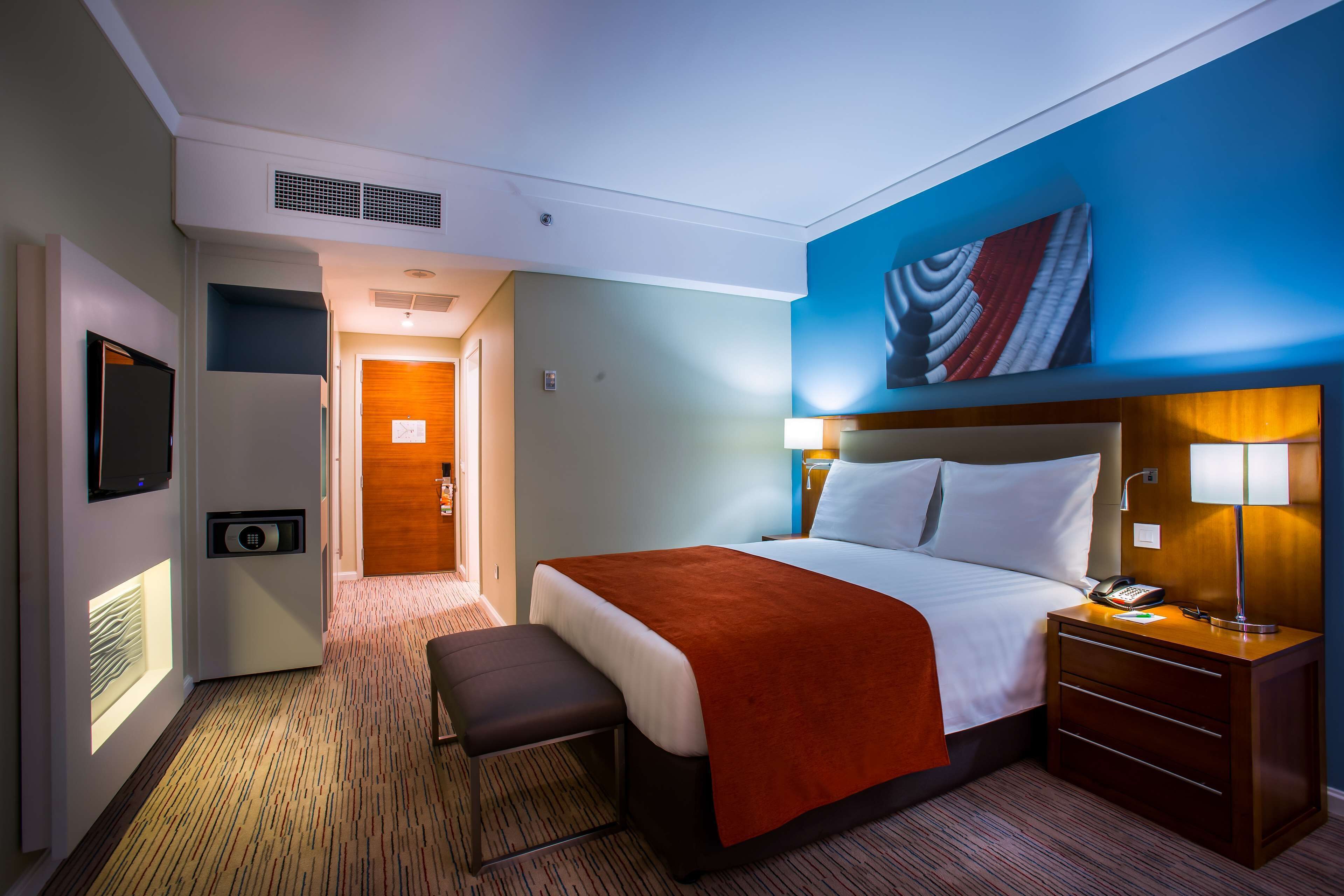 A hotel room with a bed, bedside table, lamp, and a wall-mounted artwork.
