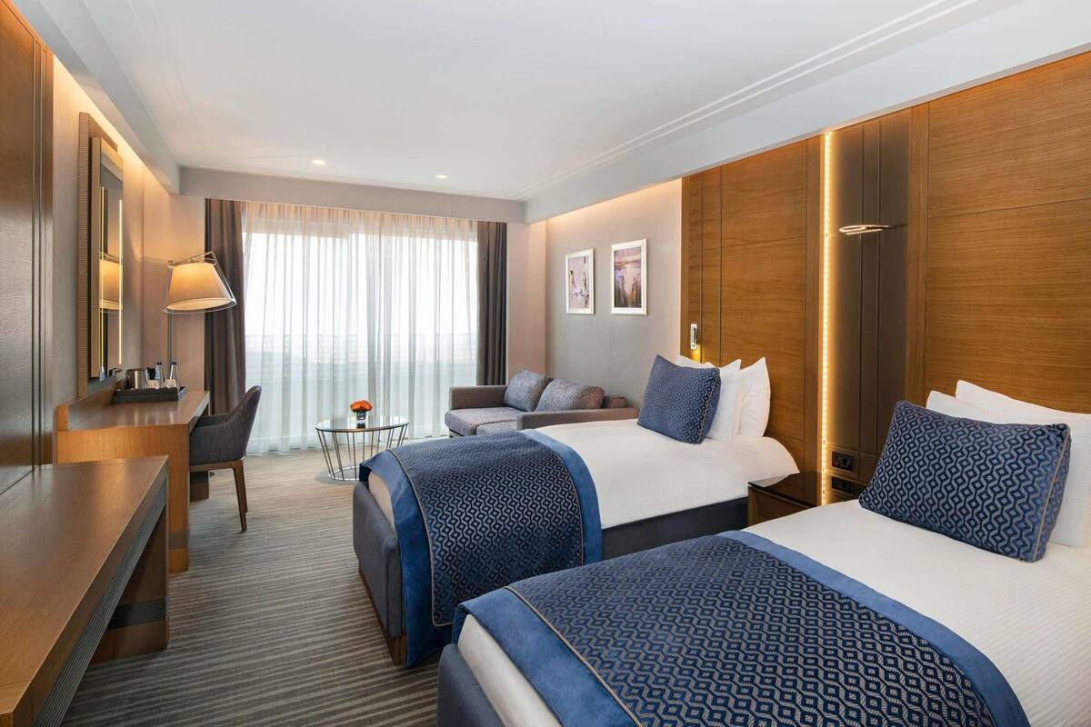 A hotel room with two beds, a desk, a chair, a small table, and a large window with curtains.