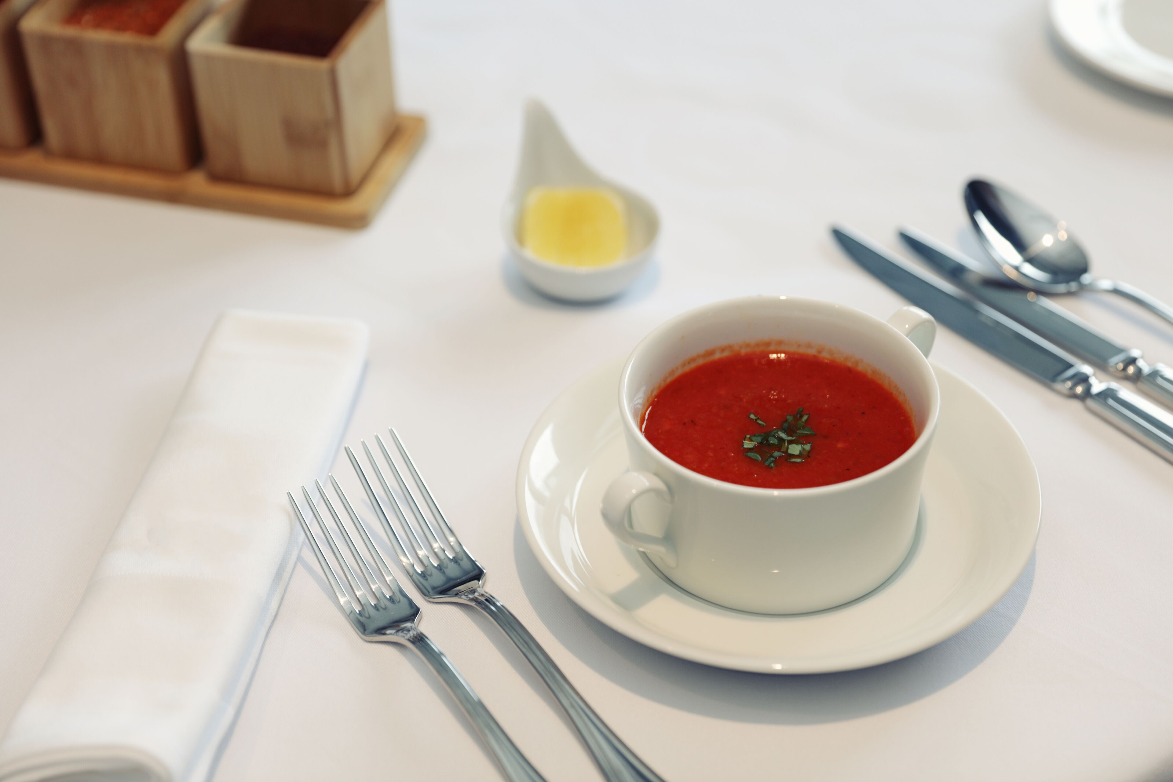 Un tavolo apparecchiato con un piatto bianco, una tazza bianca con zuppa rossa, una forchetta, un cucchiaio e un piccolo contenitore con condimento giallo.