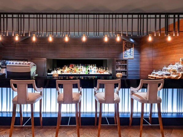 A bar area with a wooden bar counter, three bar stools, and a variety of bottles and glasses. - Mövenpick Hotel & Thermal Spa (Bursa)