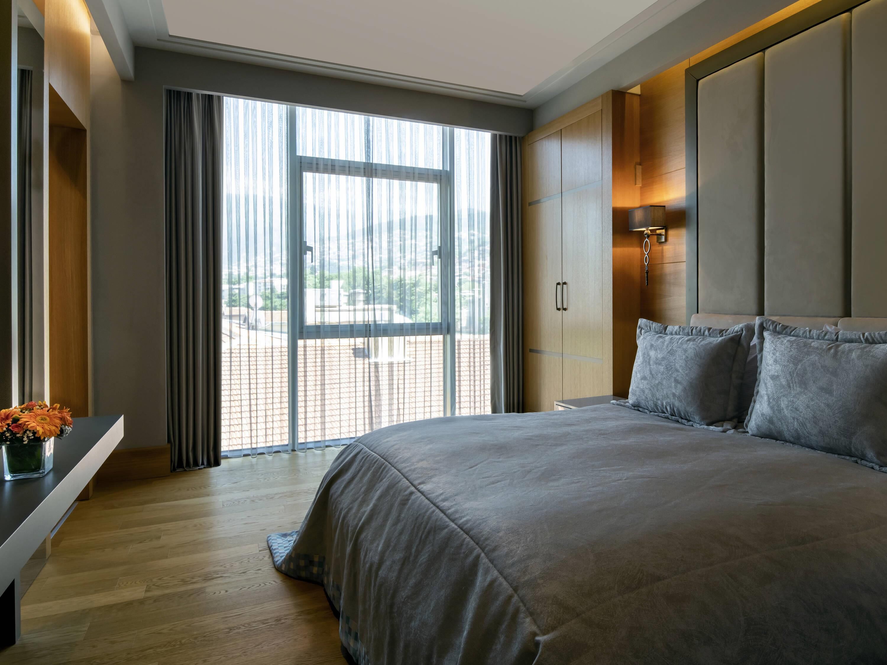 A modern hotel room with a large bed, wooden flooring, a view through a sliding door, and a small table with a vase of flowers.