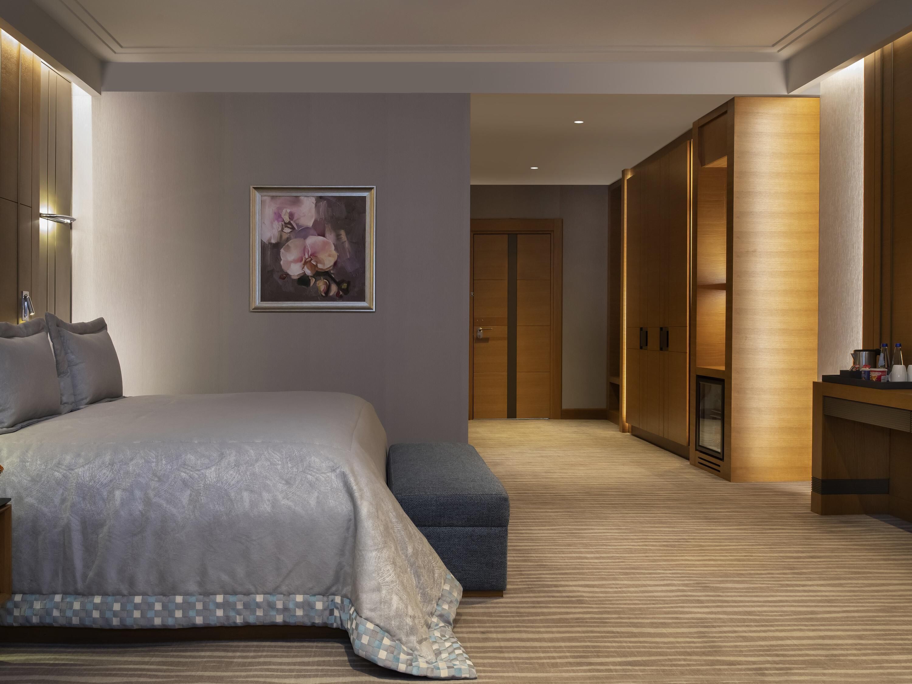 Una habitación de hotel moderna con una cama grande, un armario de madera, un pequeño rincón de descanso y un cuadro en la pared.