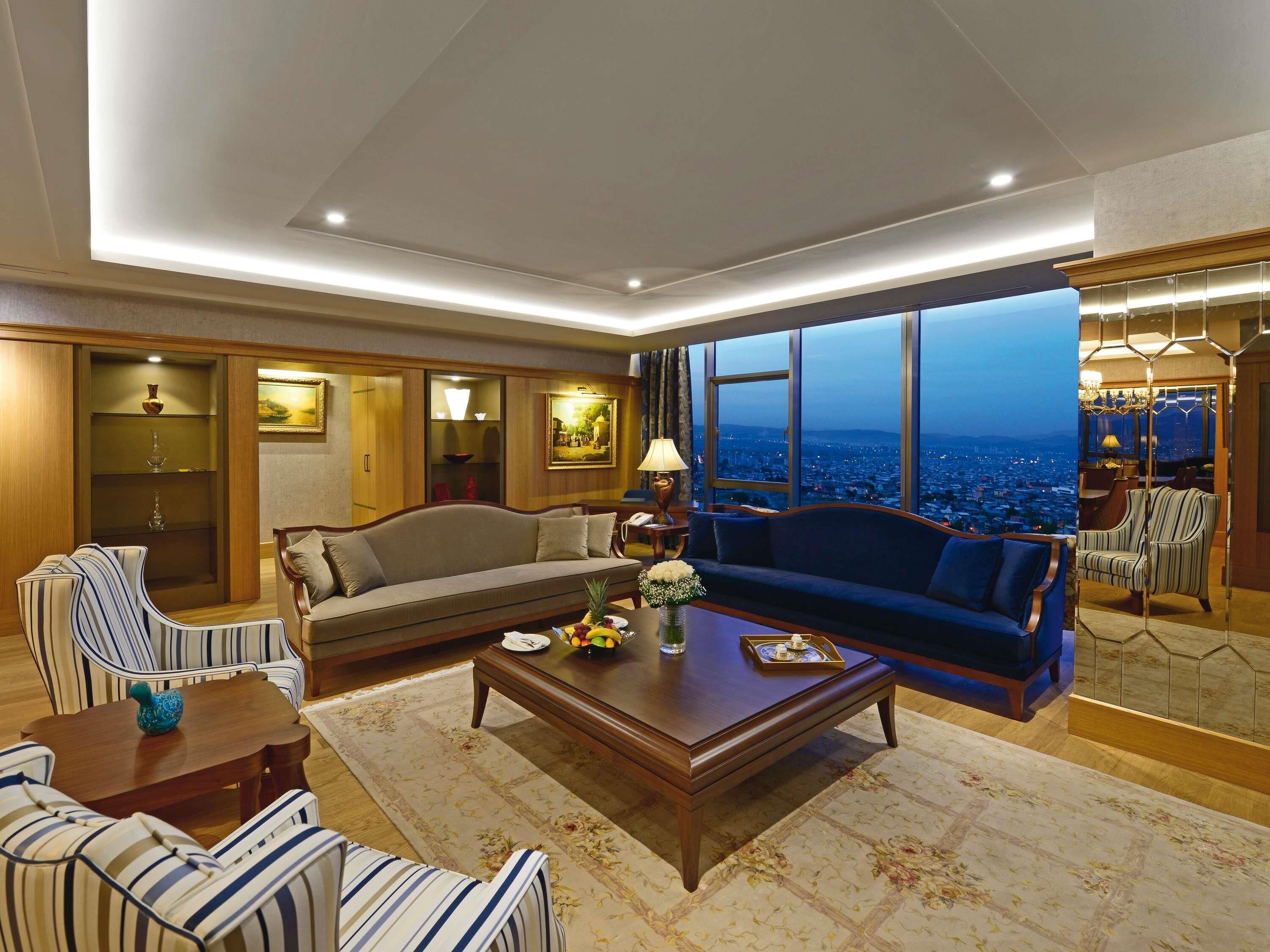 A spacious living room with a large sofa, striped chairs, a coffee table, and a view of a cityscape through large windows.