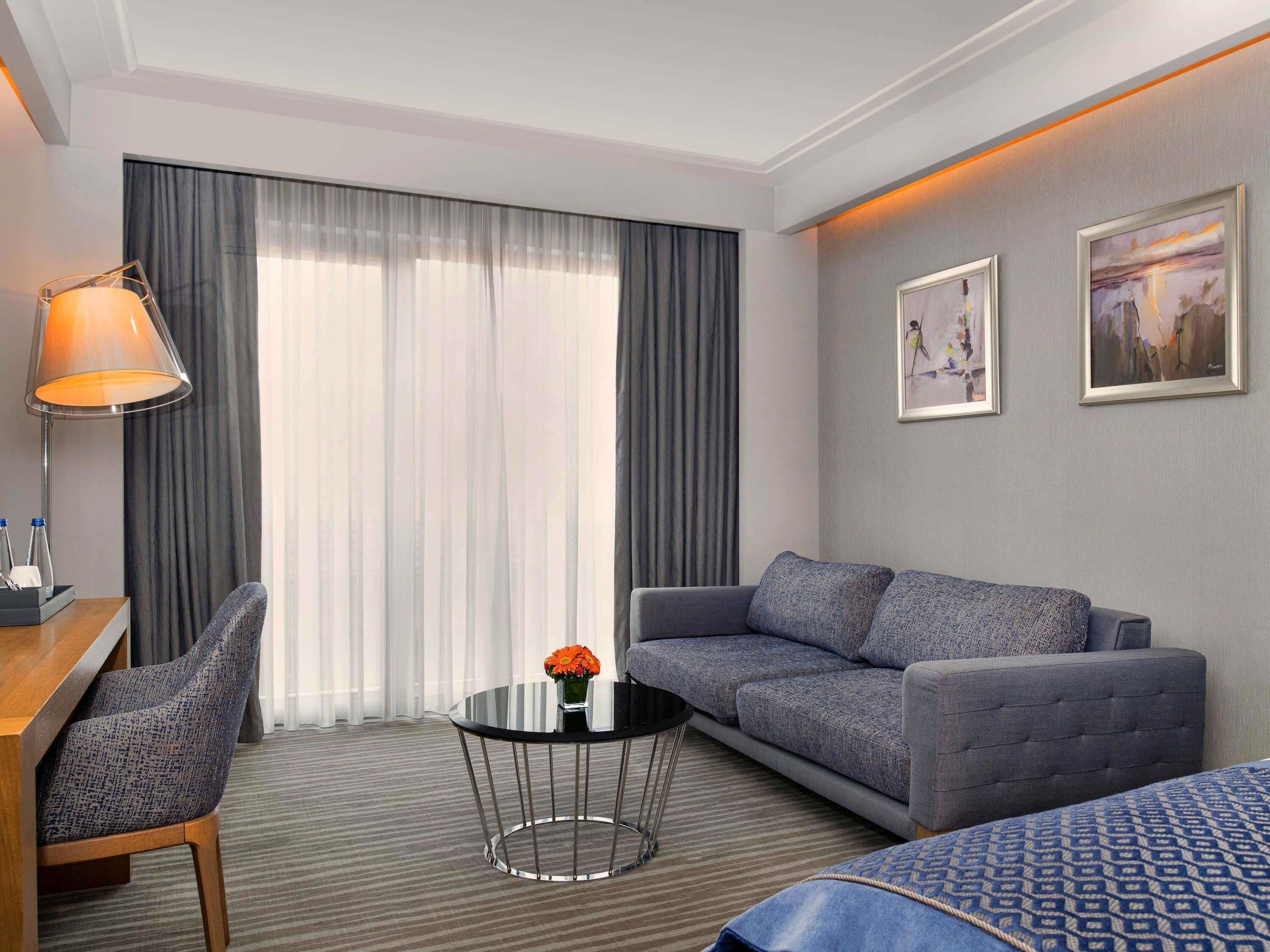A hotel room with a sofa, a small round table with a vase of flowers, a lamp, and two framed pictures on the wall.