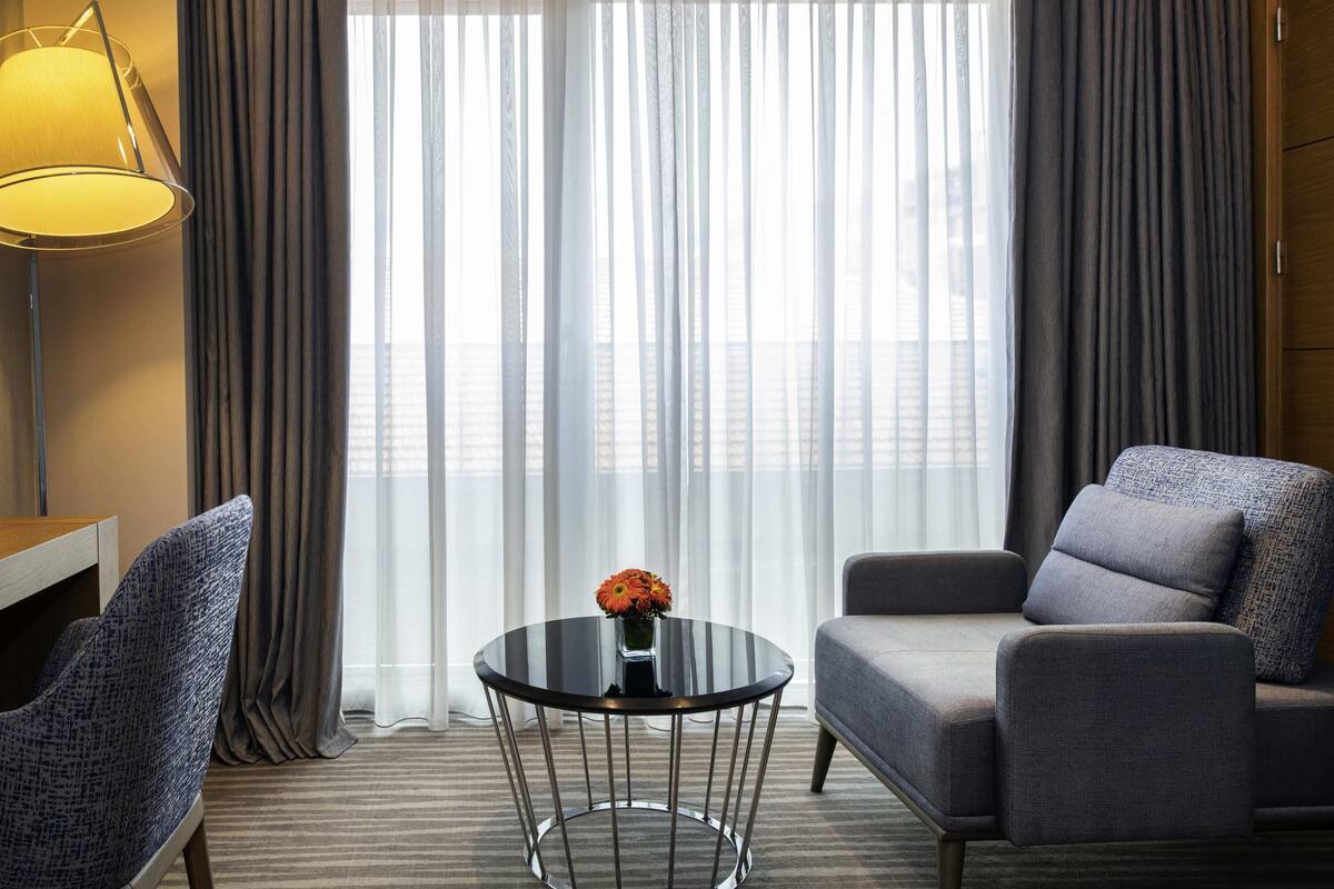 Une chambre d’hôtel avec une table ronde en verre, deux fauteuils, une lampe et une vue à travers des rideaux transparents.