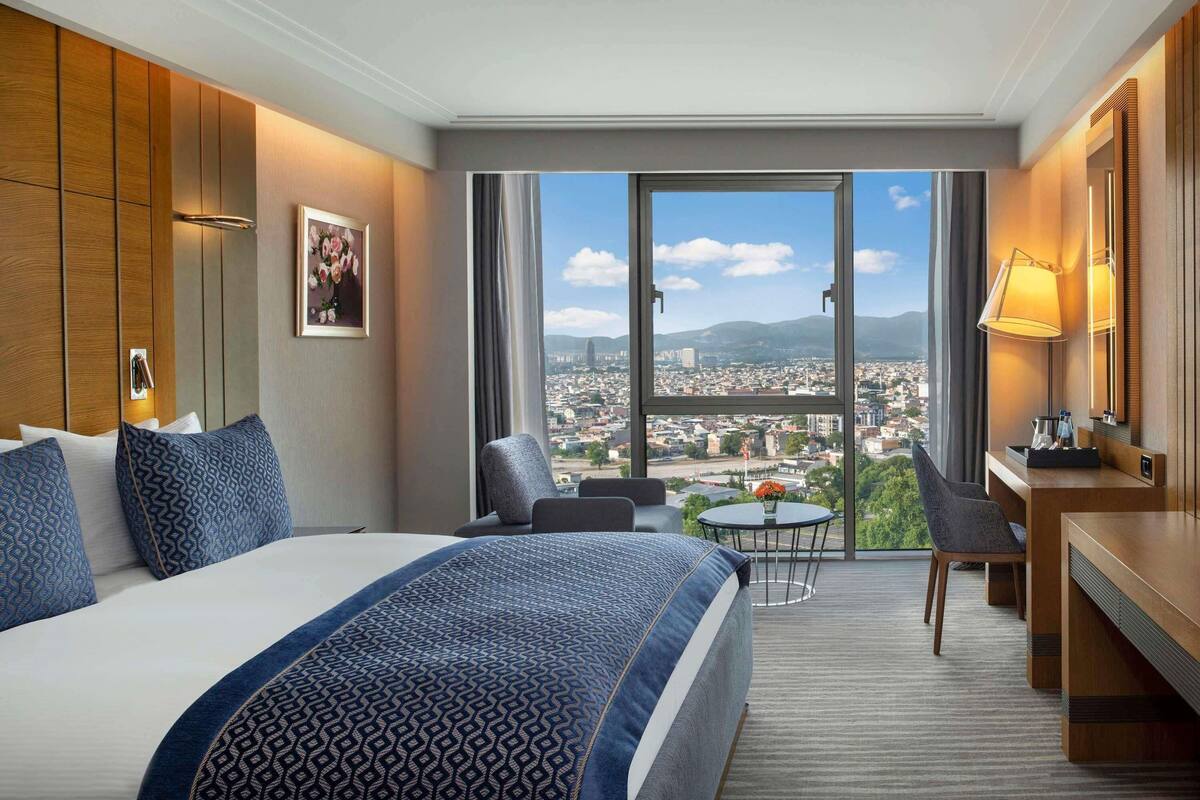 A hotel room with a large bed, a desk, a chair, and a balcony with a city view.