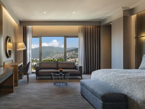 A hotel room with a large bed, a sofa, a desk, and a view of a cityscape through large windows. - Mövenpick Hotel & Thermal Spa (Bursa)
