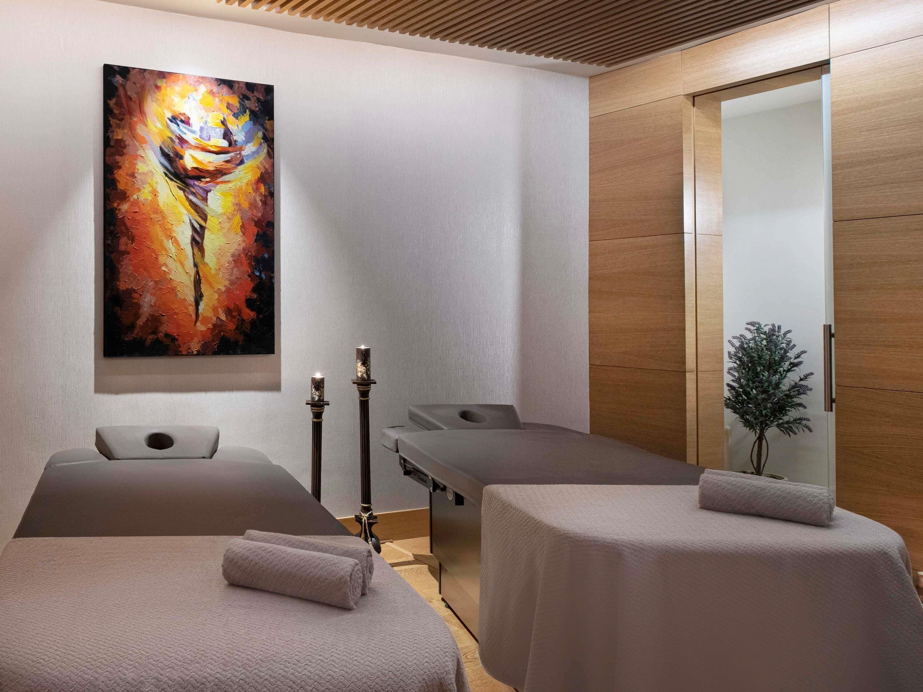 A modern spa room with two massage tables, a large abstract painting, and a wooden cabinet.