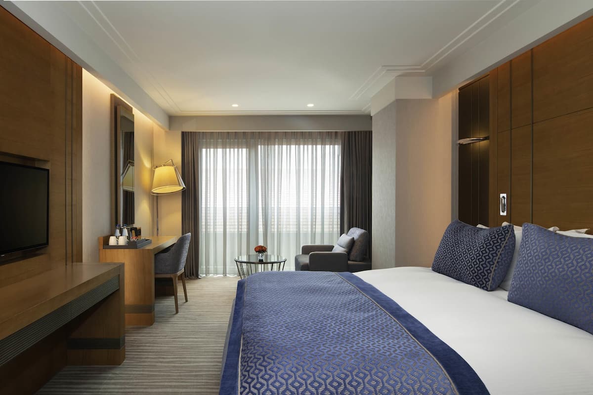 A hotel room with a large bed, a desk with a chair, a television, and a view of the city through large windows.