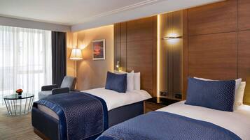 A hotel room with two beds, a desk, a chair, a lamp, and a large window.