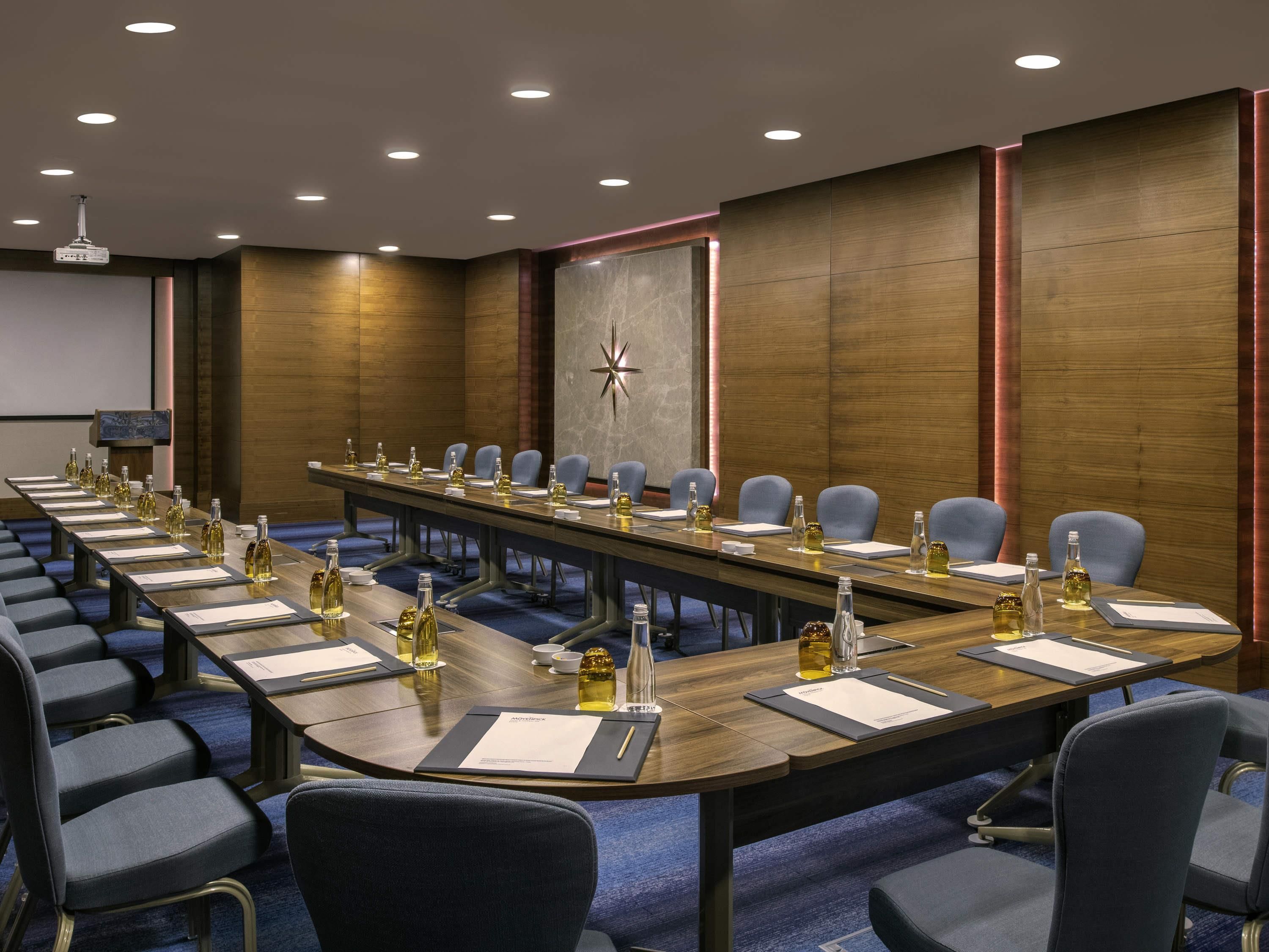 Un salón de conferencias con una larga mesa, sillas y una pantalla de proyector.