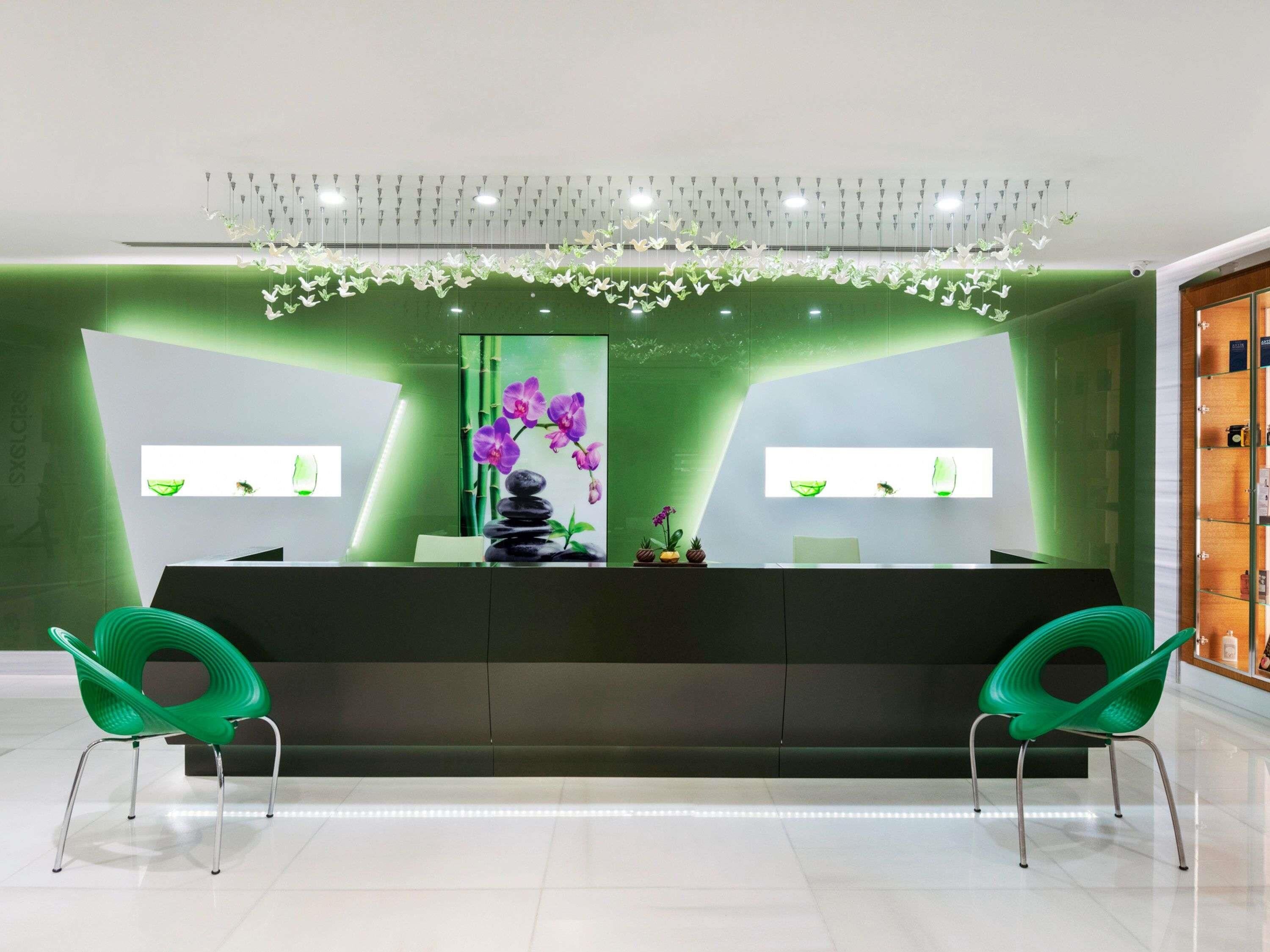 A reception area with a dark reception desk, two green chairs, a hanging light fixture, and a wall-mounted display with a stack of stones and purple orchids.