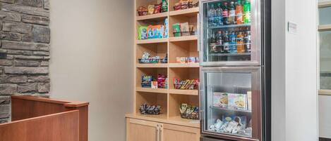 Una kitchenette con refrigerador lleno de bebidas y aperitivos, un mueble de madera con diversos alimentos, y una pared de piedra.