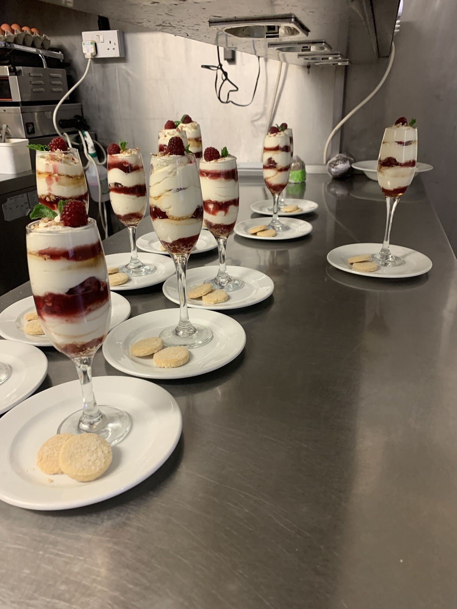 A row of dessert glasses on white plates, each topped with a strawberry and a biscuit, in a commercial kitchen setting.