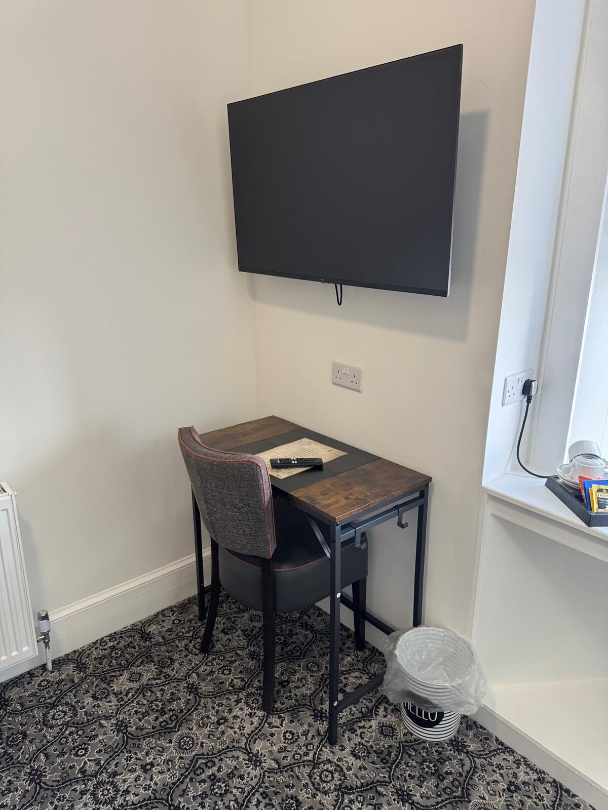 A small room with a single table, a chair, a TV mounted on the wall, and a window with a power outlet.