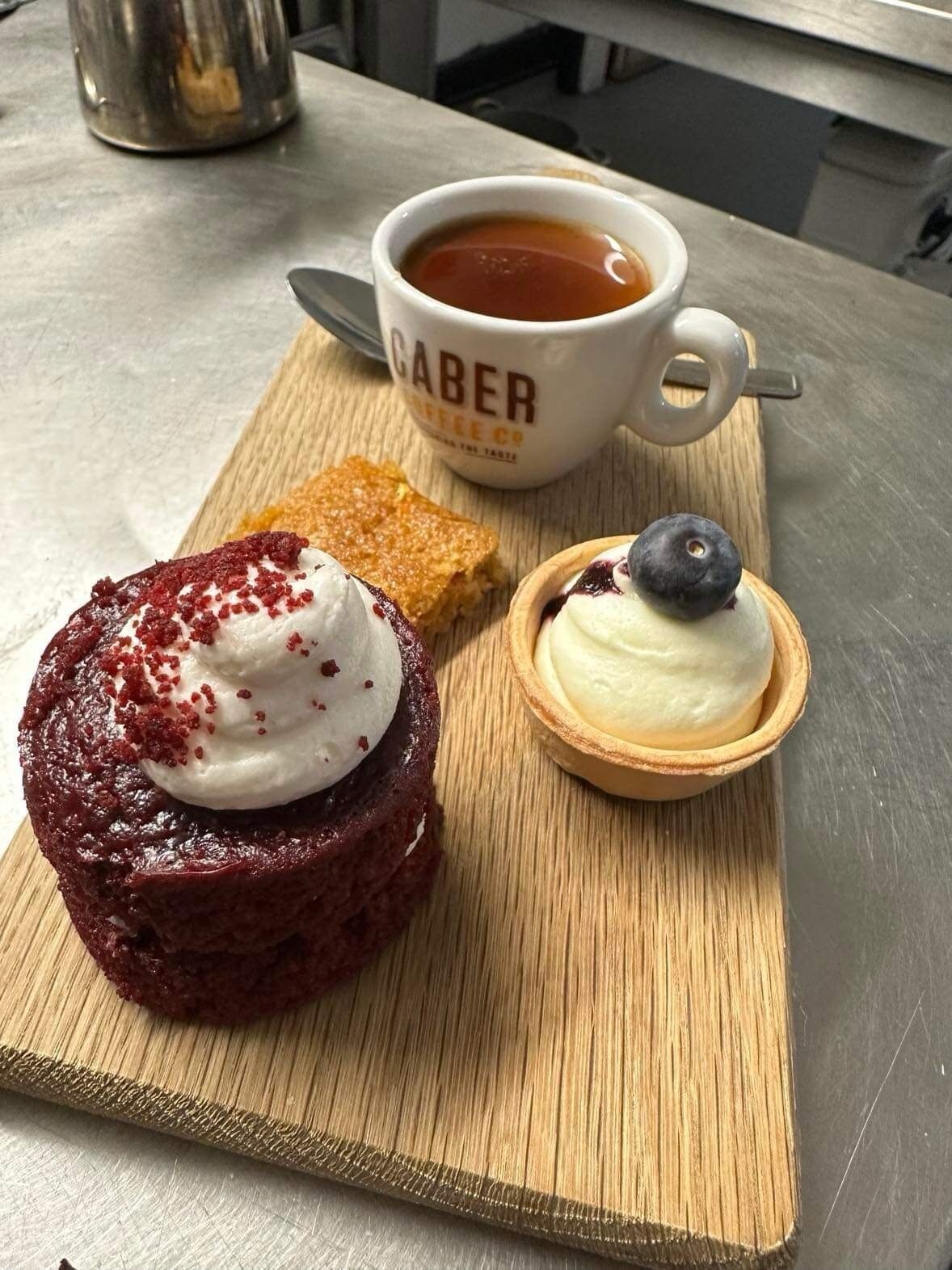 Una bandeja de postres con un pastel de terciopelo rojo cubierto de crema batida y chispas rojas, una tartaleta con arándano, y una taza de café con la etiqueta 'CABER'.