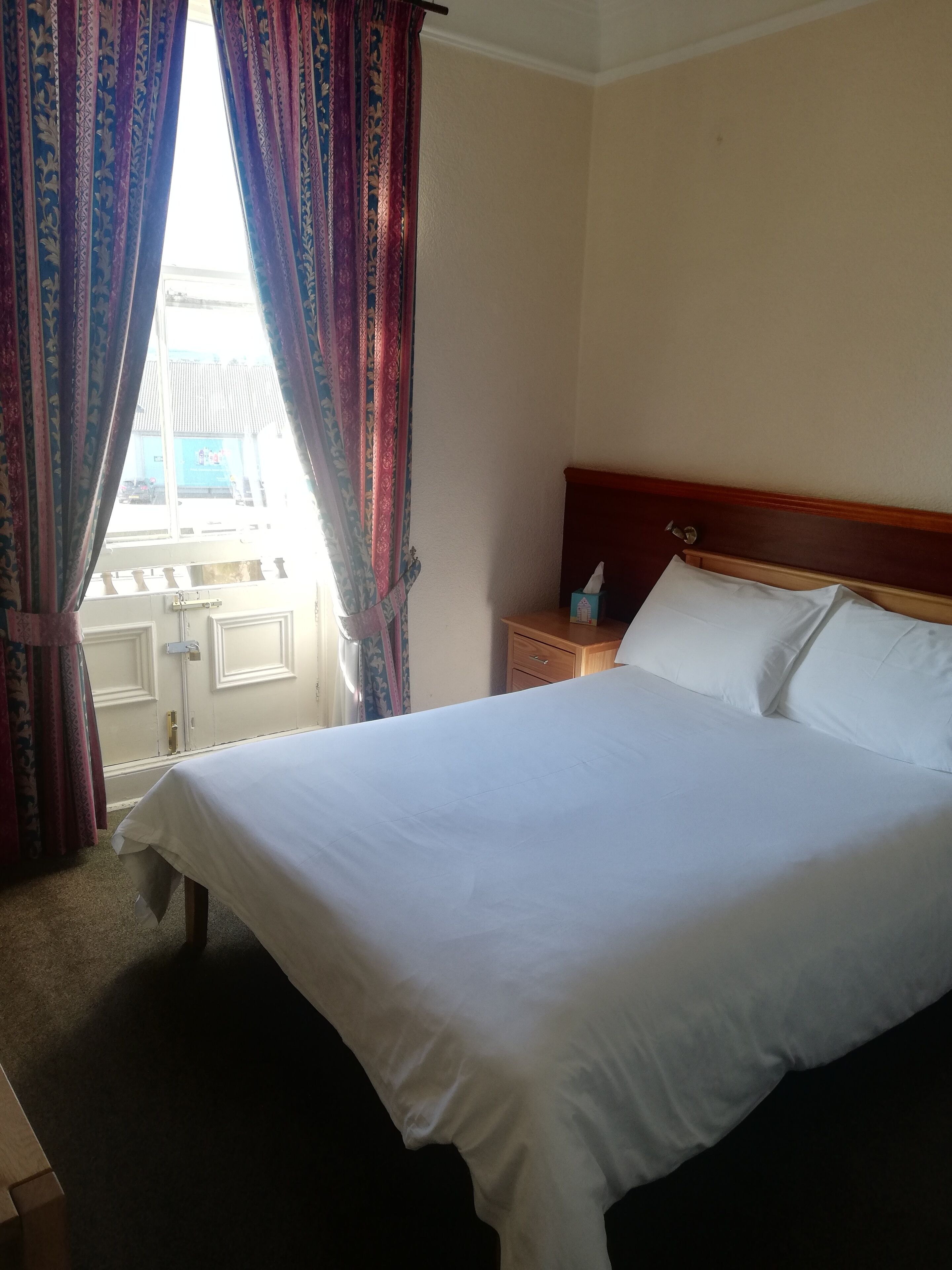 Habitación de hotel con una cama bien hecha, una mesita de noche con caja de pañuelos y vistas al exterior a través de cortinas.