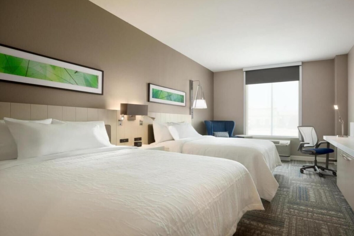 A hotel room with two beds, a desk, a chair, and a large window.