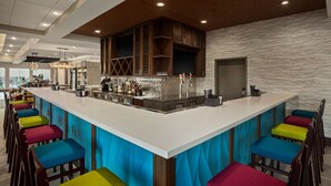Un espace bar moderne avec un long comptoir, des tabourets de bar colorés et une variété de bouteilles et de verres.