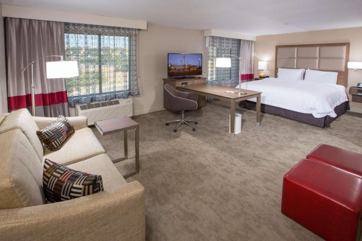 A hotel room with a bed, a desk, a chair, a TV, and a red ottoman.