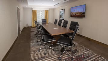 A conference room with a long table, multiple chairs, a carpeted floor, a television on the wall, and a door on the left.