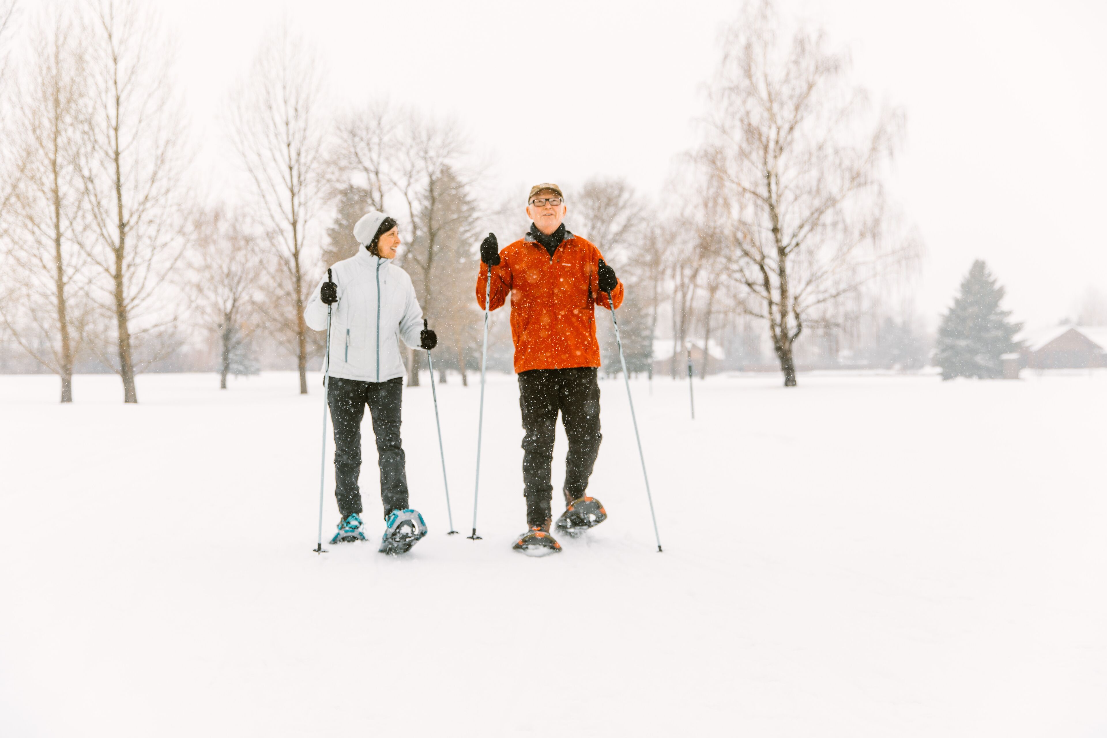 Two people cross-country skiing in a snowy landscape with bare trees and a house in the background.