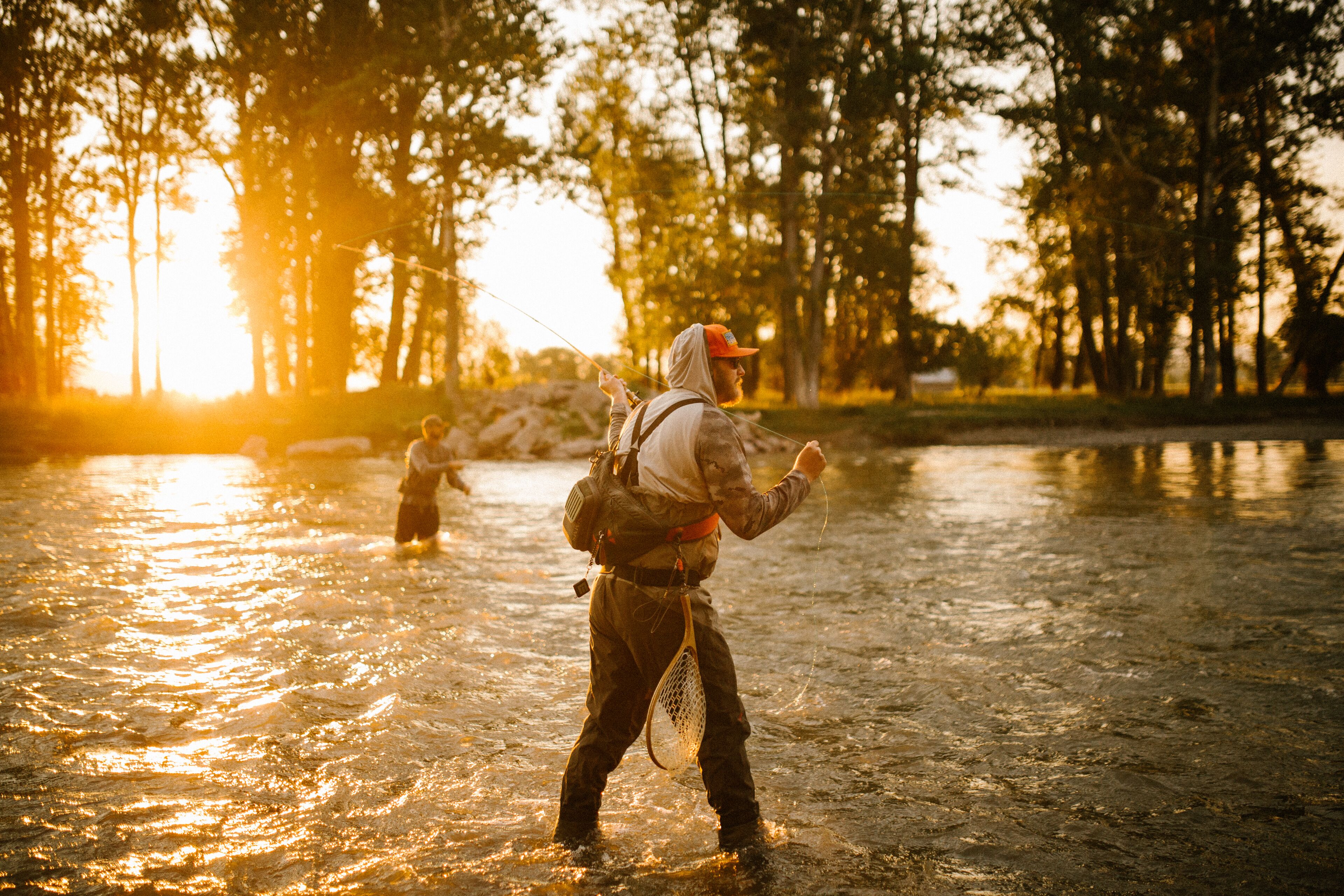Una persona de pie en un río con una caña de pescar, rodeada de árboles y luz solar.