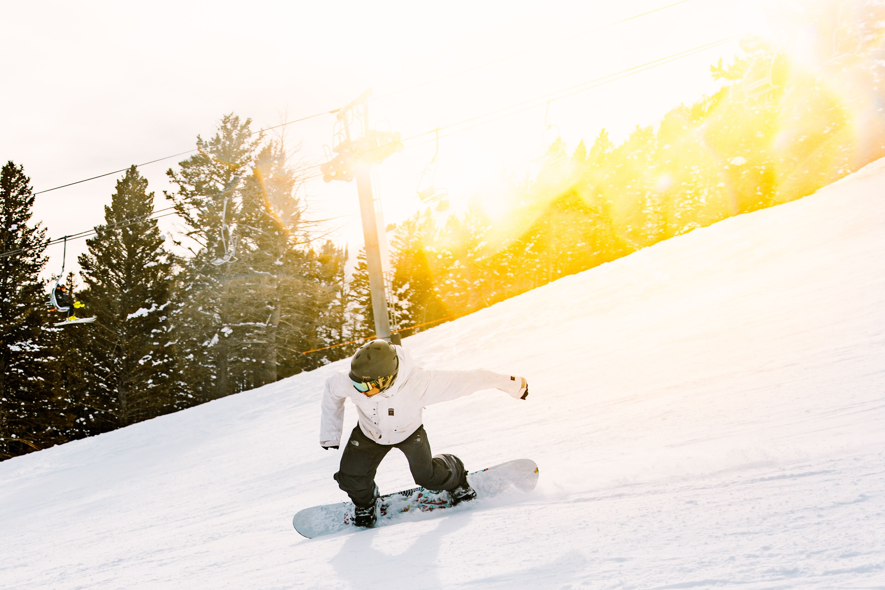Un snowboarder en una ladera nevada con árboles y un telesilla al fondo.