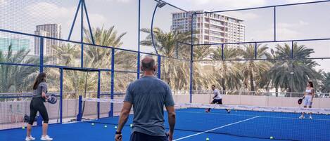 An outdoor tennis court with a net, surrounded by a blue fence and palm trees.