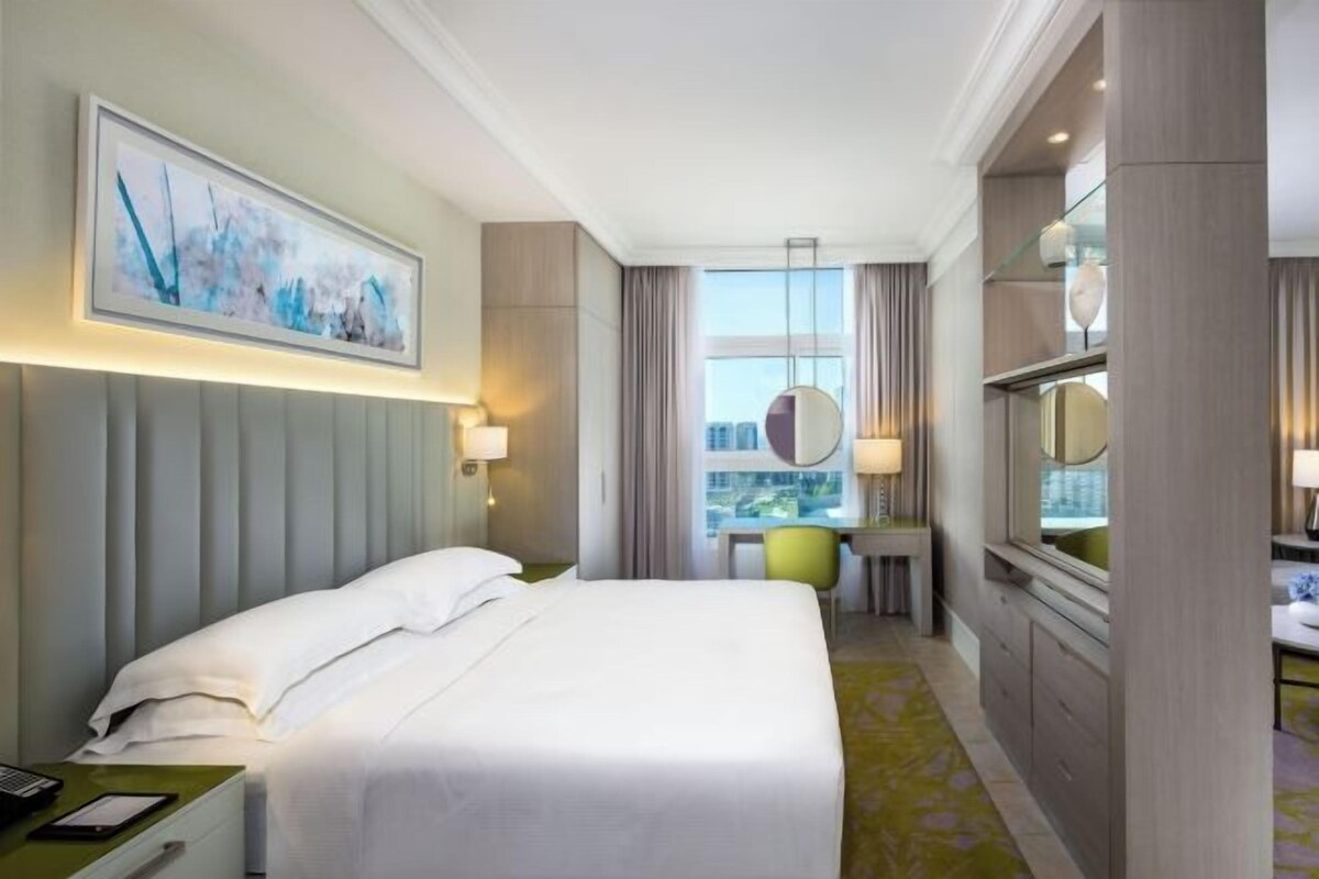 Quarto de hotel com cama, mesa de cabeceira, luminária, janela e vista para a cidade.