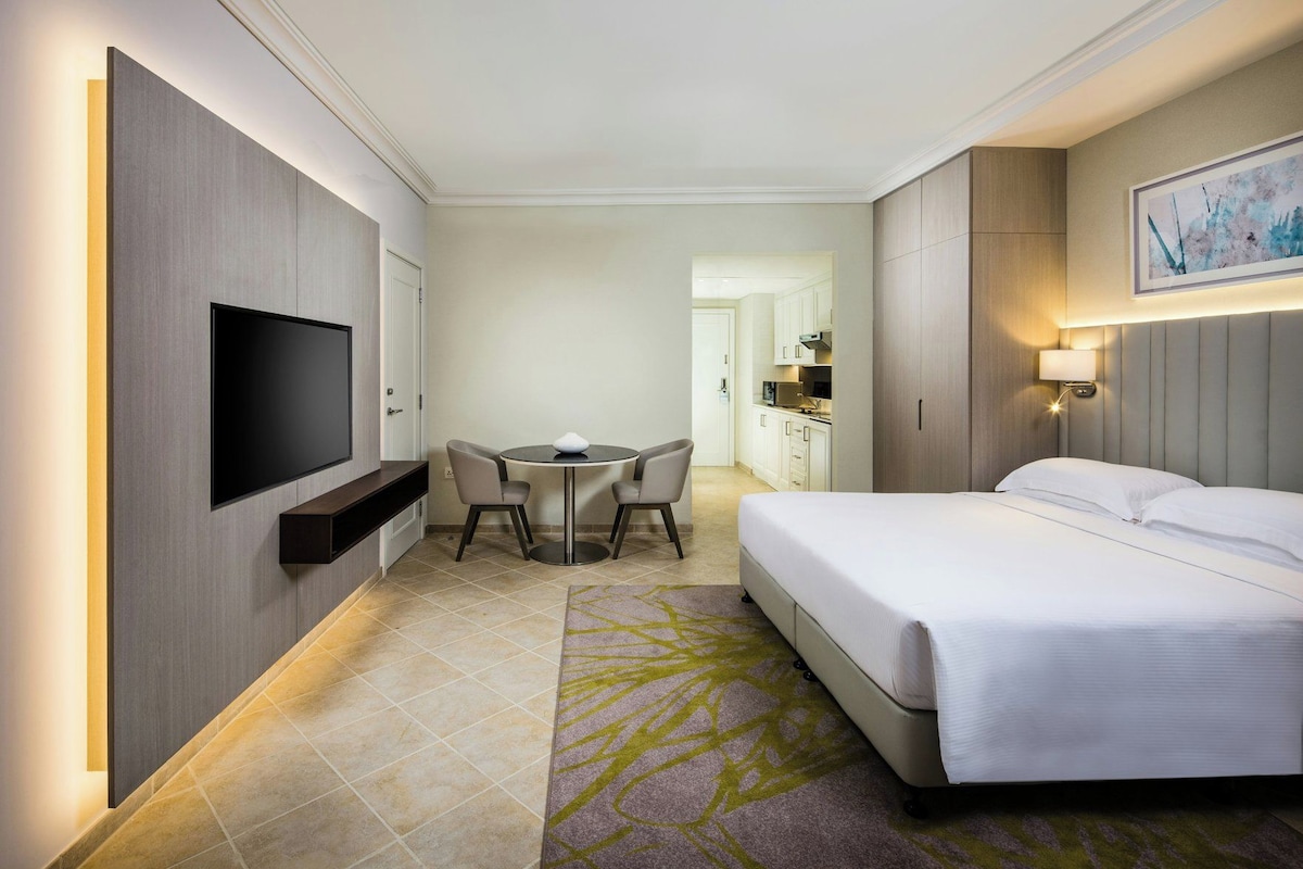 A hotel room with a bed, a TV mounted on the wall, a dining table with chairs, and a kitchenette in the background.