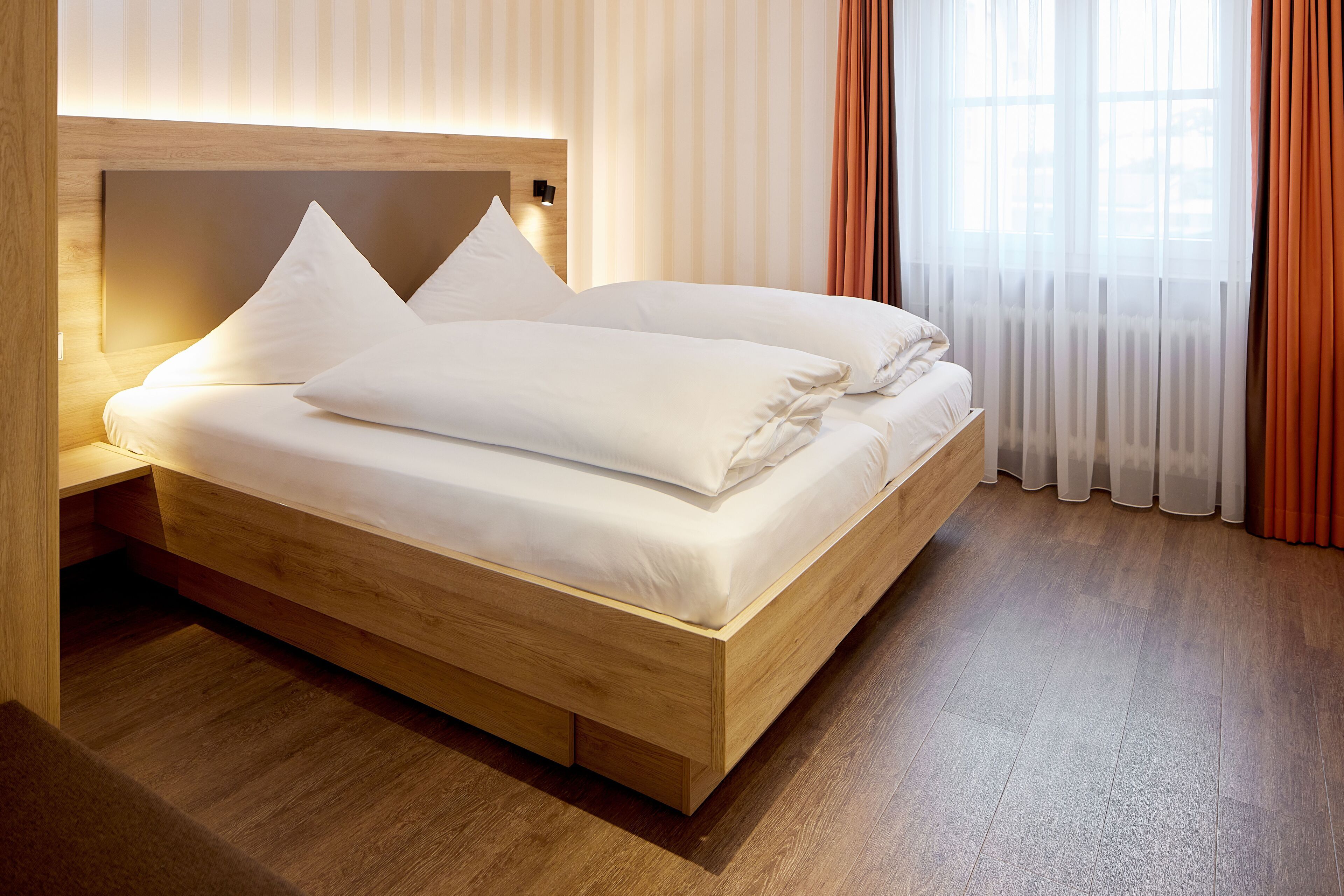 Una habitación de hotel moderna con una cama de madera, una mesita de noche y un ventanal con cortinas translúcidas.
