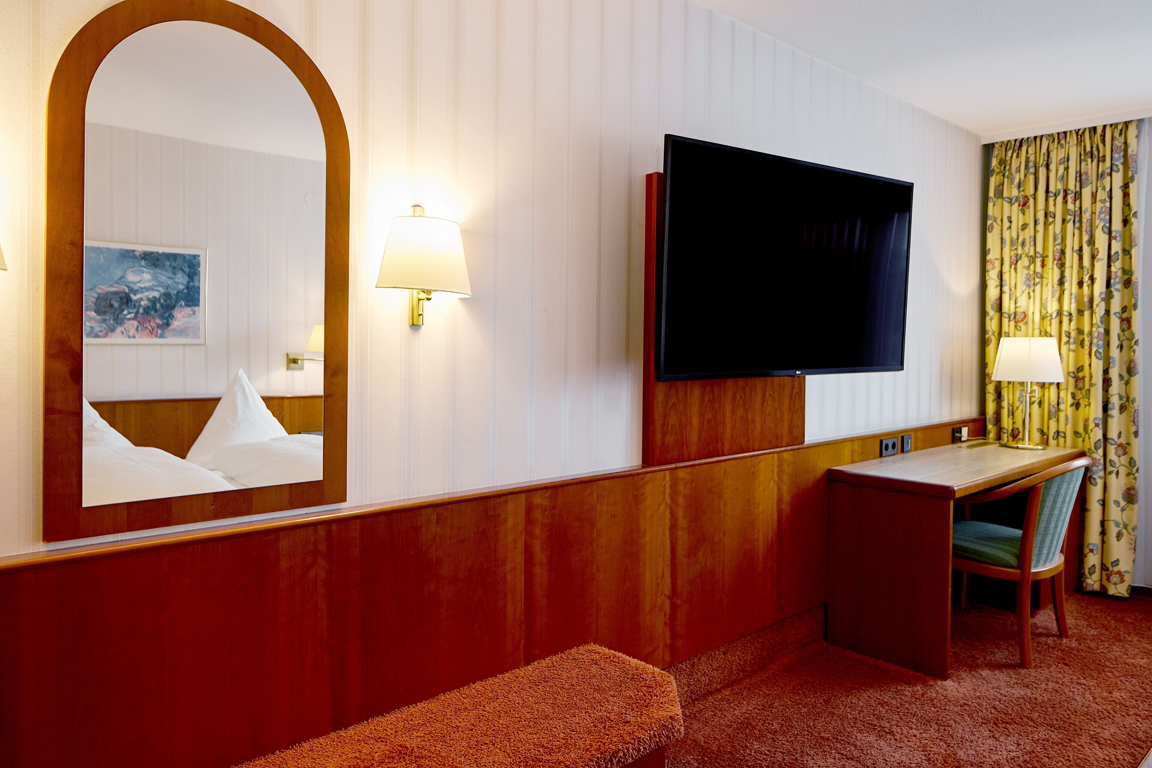 A hotel room with a large mirror, a flat-screen TV, a desk with a chair, and a bench.