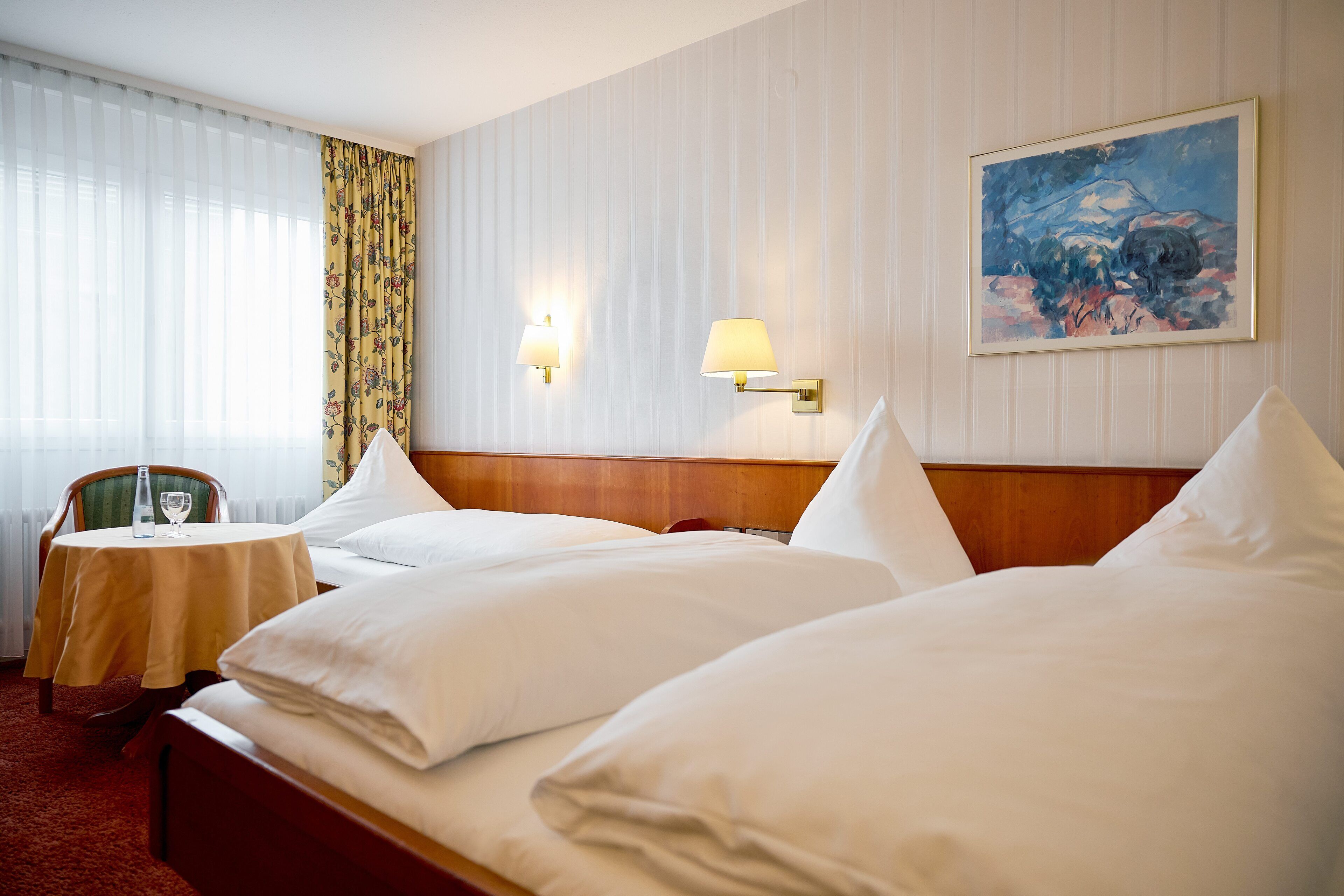 A hotel room with two beds, a small table with a chair, a painting on the wall, and a window with curtains.