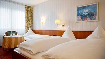 A hotel room with two beds, a small table with a chair, a painting on the wall, and a window with curtains.