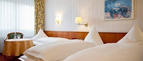 A hotel room with two beds, a small table with a chair, a painting on the wall, and a window with curtains.