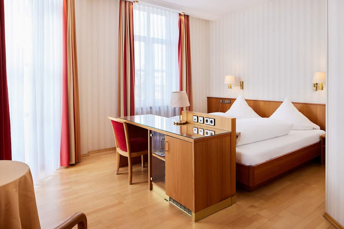Habitación de hotel con escritorio de madera, cama con sábanas blancas, silla roja y ventana grande con cortinas.