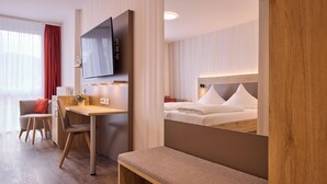A hotel room with a wooden desk, a television mounted on the wall, a bed with white linens, and a bench.