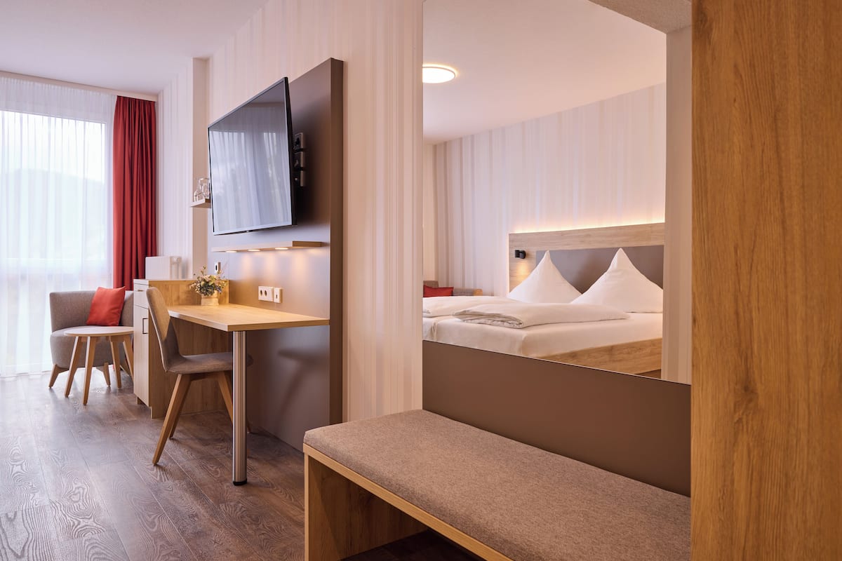 Habitación de hotel con un escritorio de madera, un televisor montado en la pared, una cama con ropa blanca y un banco.