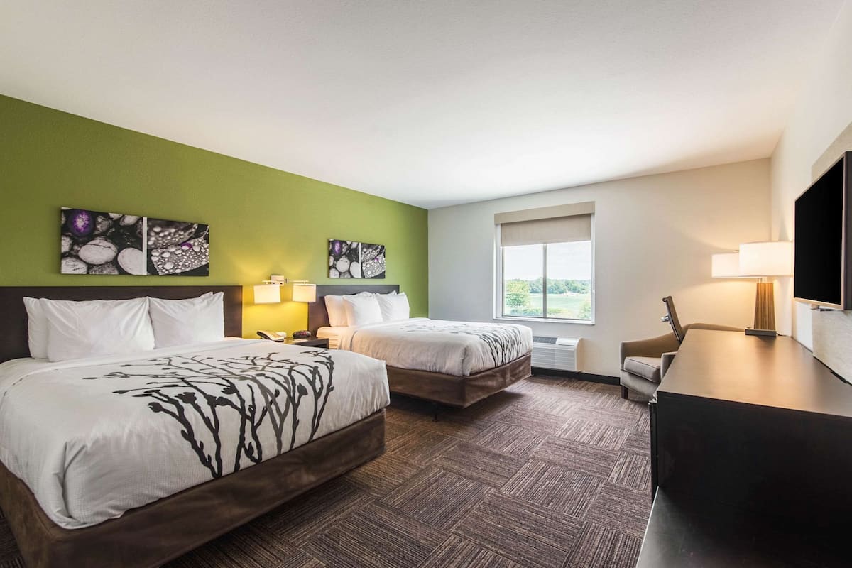 Ein Hotelzimmer mit zwei Betten, einem Schreibtisch und einem Fernseher. An den Wänden hängen zwei Bilder und ein Fenster bietet Blick ins Freie.