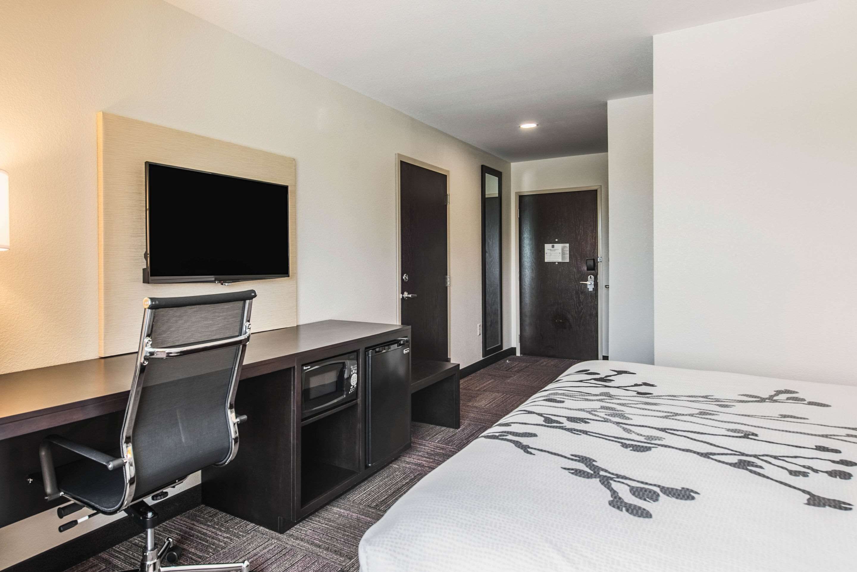 Quarto de hotel com uma cama, uma escrivaninha com cadeira, televisão e porta.