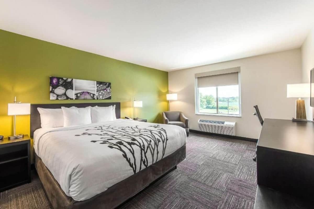 Ein Hotelzimmer mit einem Bett, zwei Nachttischen, einem Sessel, einem Schreibtisch, einem Fenster und Bildern an der Wand.