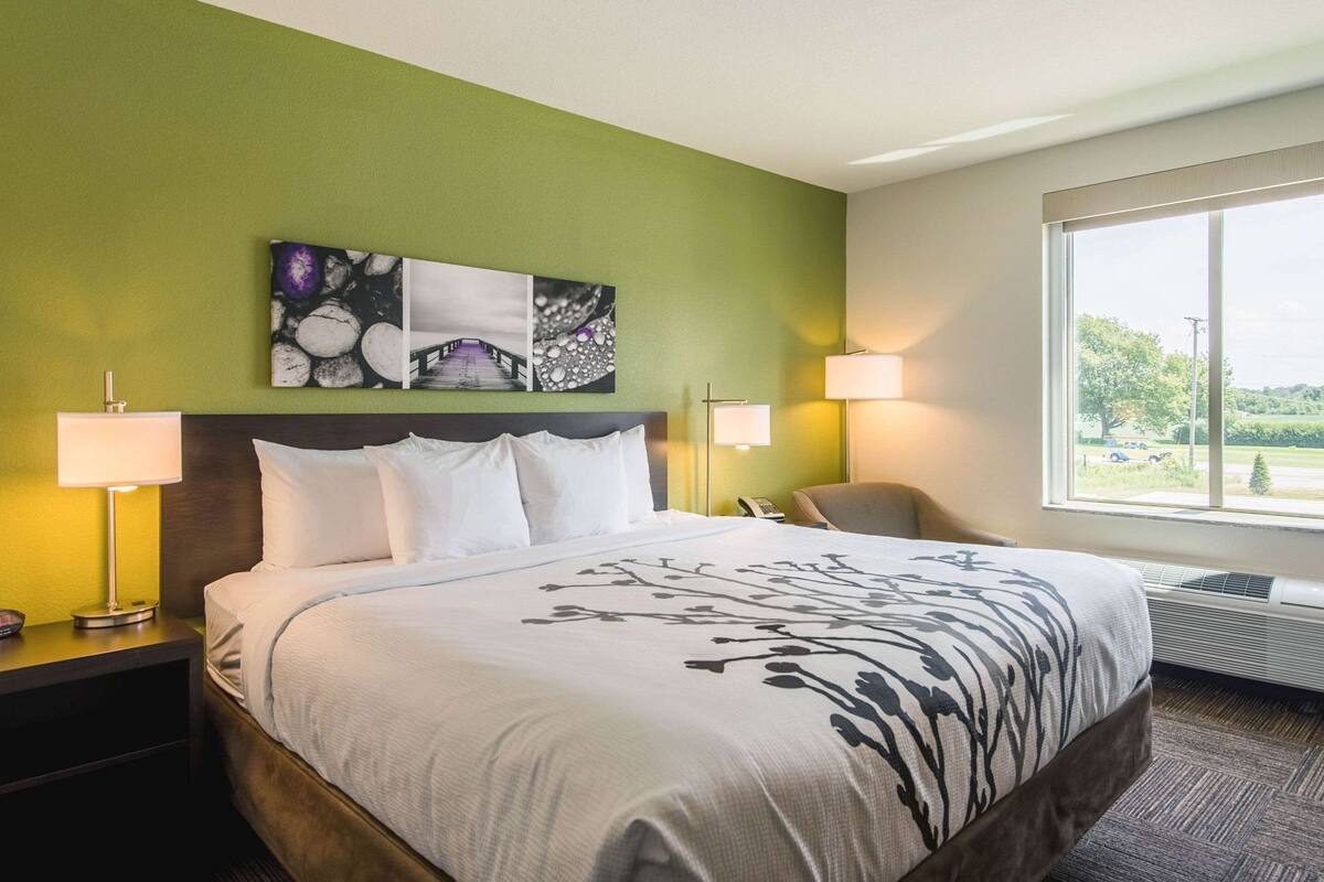 Ein Hotelzimmer mit einem großen Bett, zwei Nachttischlampen, einem Sessel, einem Fenster mit Blick und Kunstwerken an der Wand.