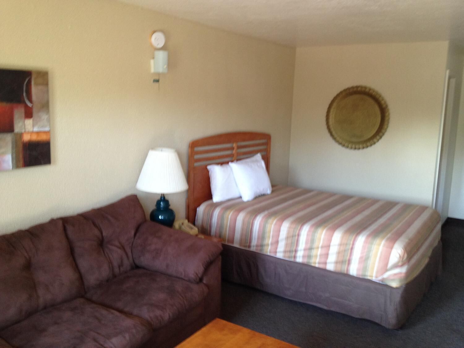 Ein Hotelzimmer mit einem braunen Sofa, einem Einzelbett mit gestreifter Bettwäsche, einem Holzkopfende, einer Lampe und einer Wanduhr.