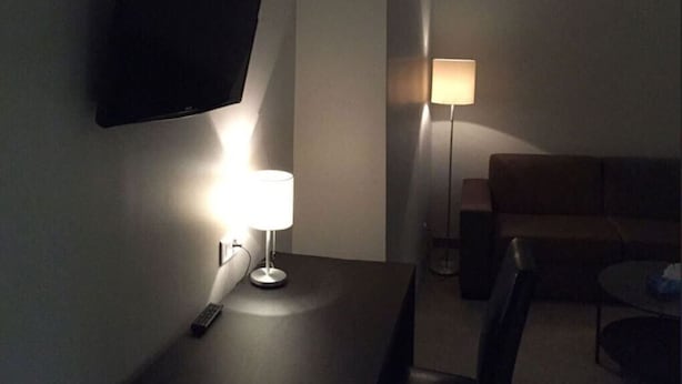 Een kamer met gedempt licht, een aan de muur bevestigde televisie, een tafel met lamp, een bank en een klein rond tafeltje.
